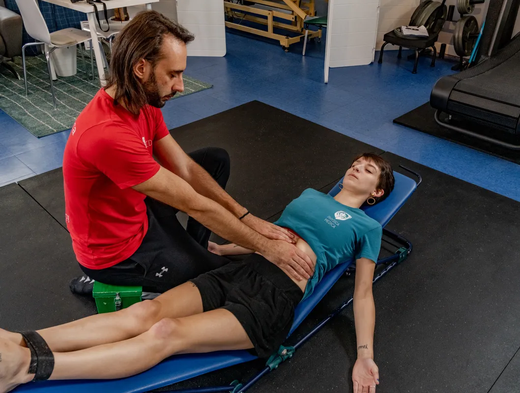 A practitioner in red branded shirt performs manual therapy on a male client's leg while the client lies on a treatment table.